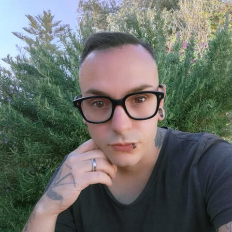 Alessi Schreiber Alessi Schreiber, with short hair and glasses poses for a selfie outdoors, surrounded by greenery. They have a tattoo on their arm and are wearing a simple t-shirt. The expression is thoughtful, with one hand resting on their face.